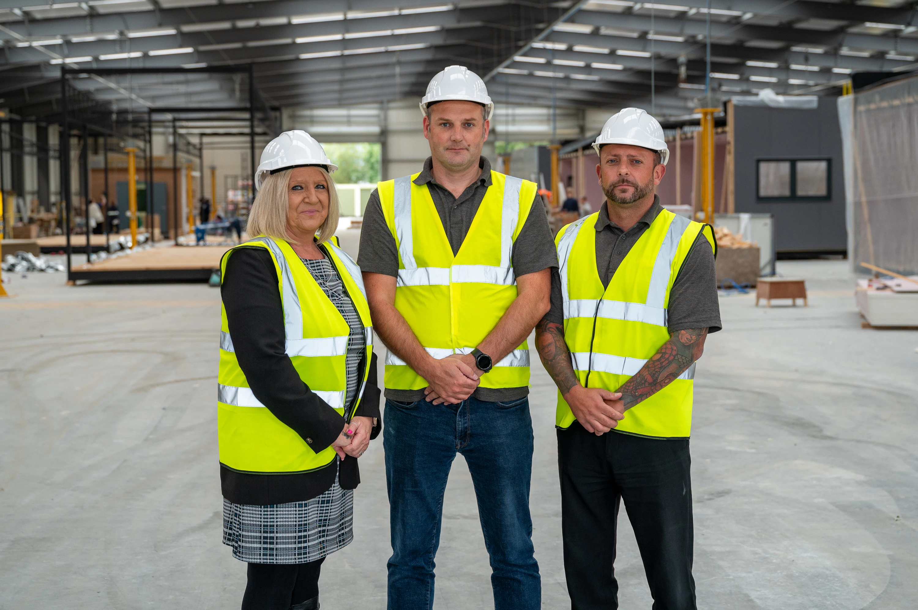 Three people, wearing white hard hats and yellow reflective vests, stand side by side inside a large industrial warehouse under construction.