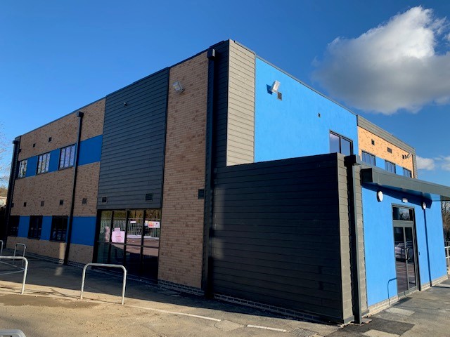 A rectangular, two-story building features a mix of brown brick and blue-panel walls with large glass windows, situated on a paved street under a clear, blue sky with scattered clouds.