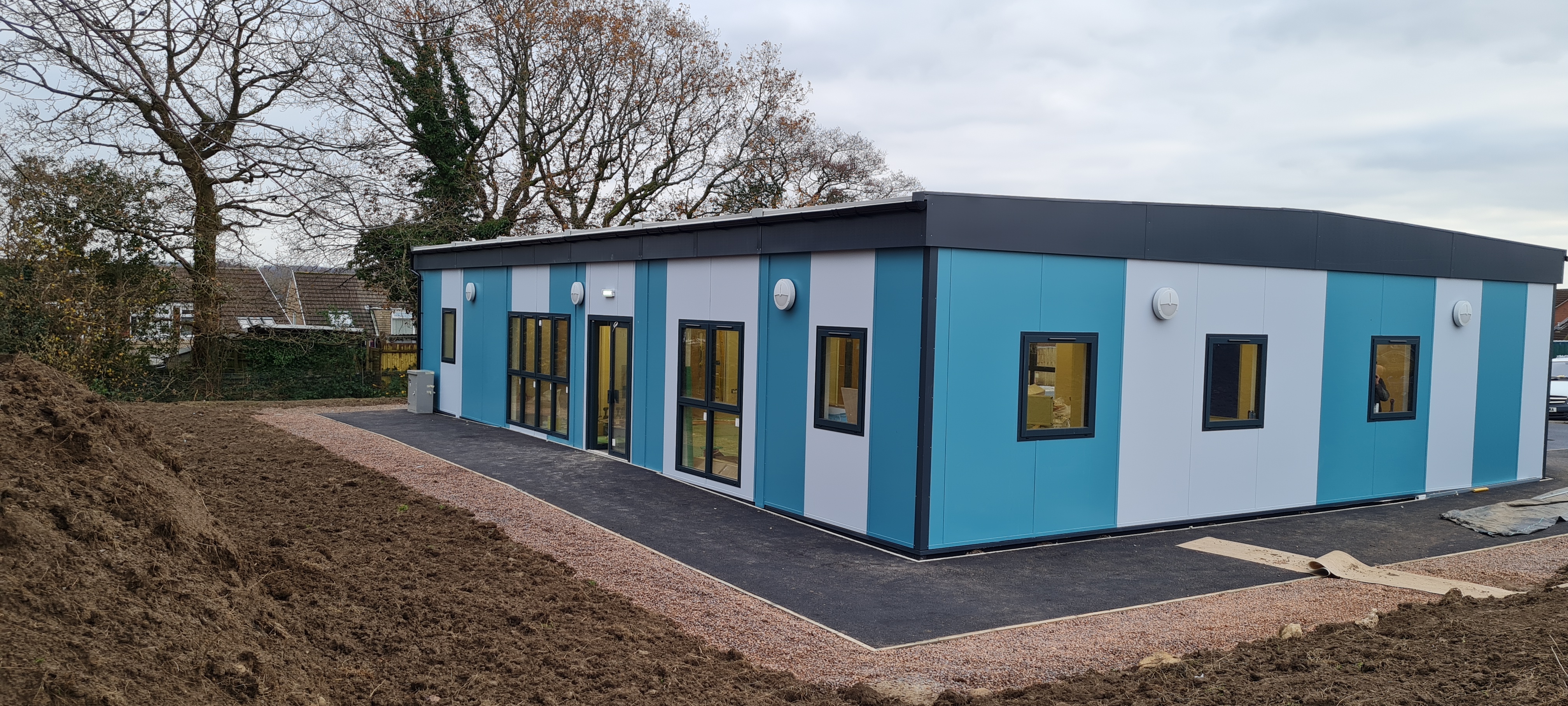 Rectangular modular building with teal and gray panels; situated on a newly prepared plot encircled by a construction path. Leafless trees and residential homes are visible in the background.