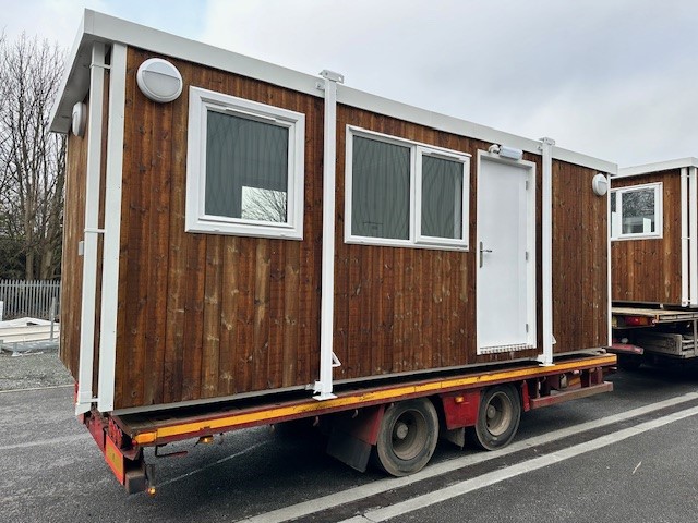 A brown wooden portable cabin with white-framed windows and a door is being transported on a red flatbed trailer on a road next to another similar cabin.