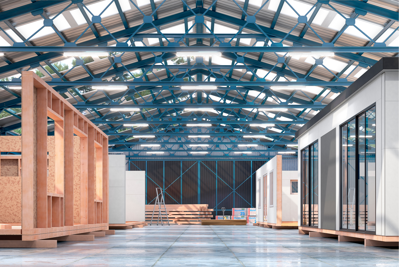 Modules under construction in a spacious warehouse, with wooden frames and finished segments on the floor; steel blue beams and roof lights above, pallets and ladders scattered around.