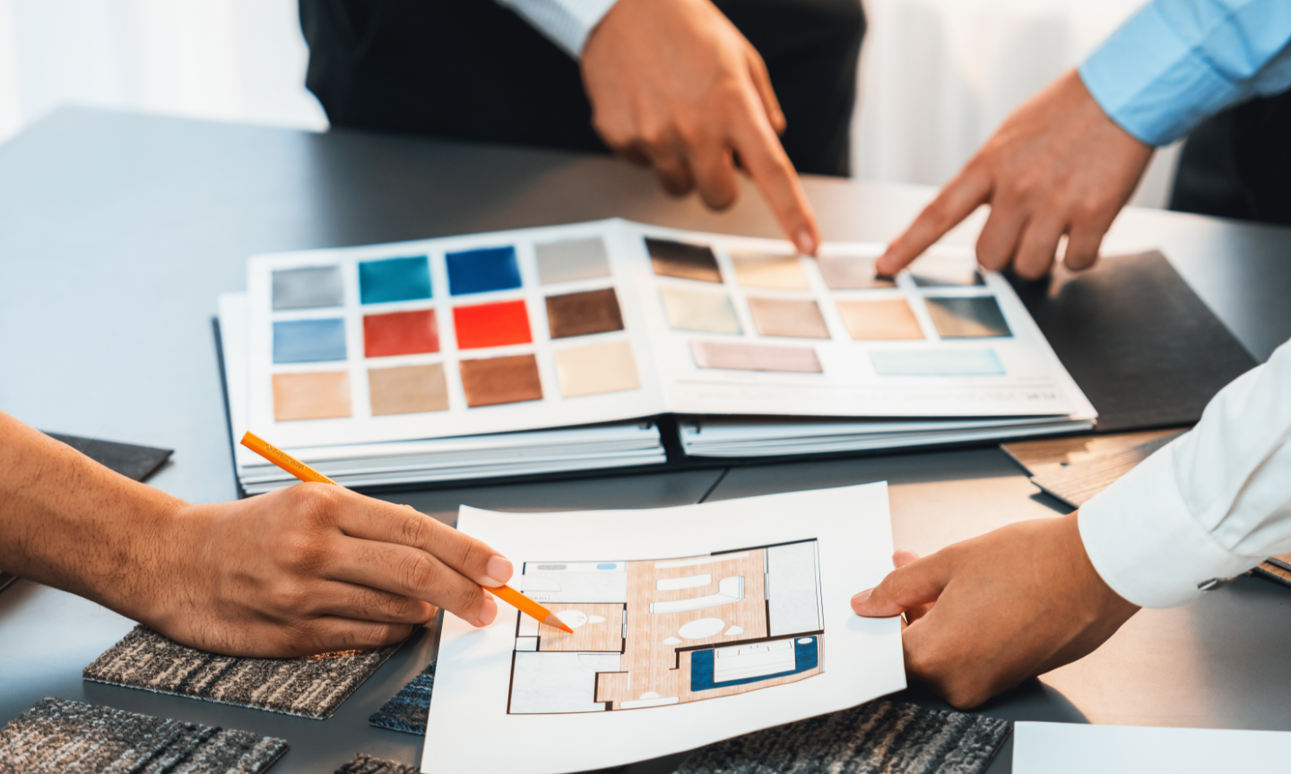 Three hands pointing at a table-top selection of color swatches and a floor plan, engaged in a decision-making process in a well-lit office setting.