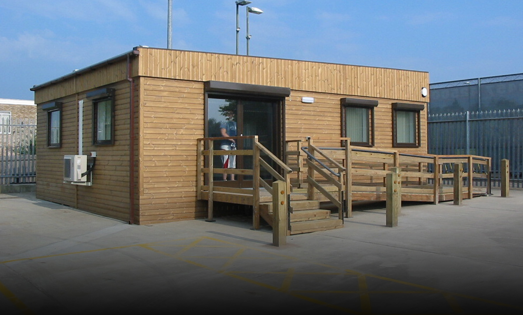 A small wooden modular building with windows, a door, air conditioning unit, and a ramp/staircase entrance, situated in a fenced industrial area under a clear sky.
