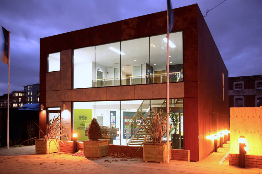 A modern, two-story glass-walled building with illuminated interior and exterior lights sits on a paved area at dusk, with potted plants outside and visible staircase inside.