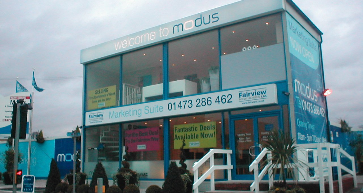 A two-story marketing suite with large windows displays signs reading "welcome to modus," "fantastic deals available now!" and "Fairview New Homes Ltd.," with a contact number 01473 286 462.