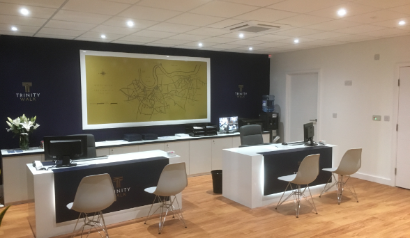 An office reception area with two desks featuring computers and chairs, set against a wall with a large gold map and "Trinity Walk" branding.