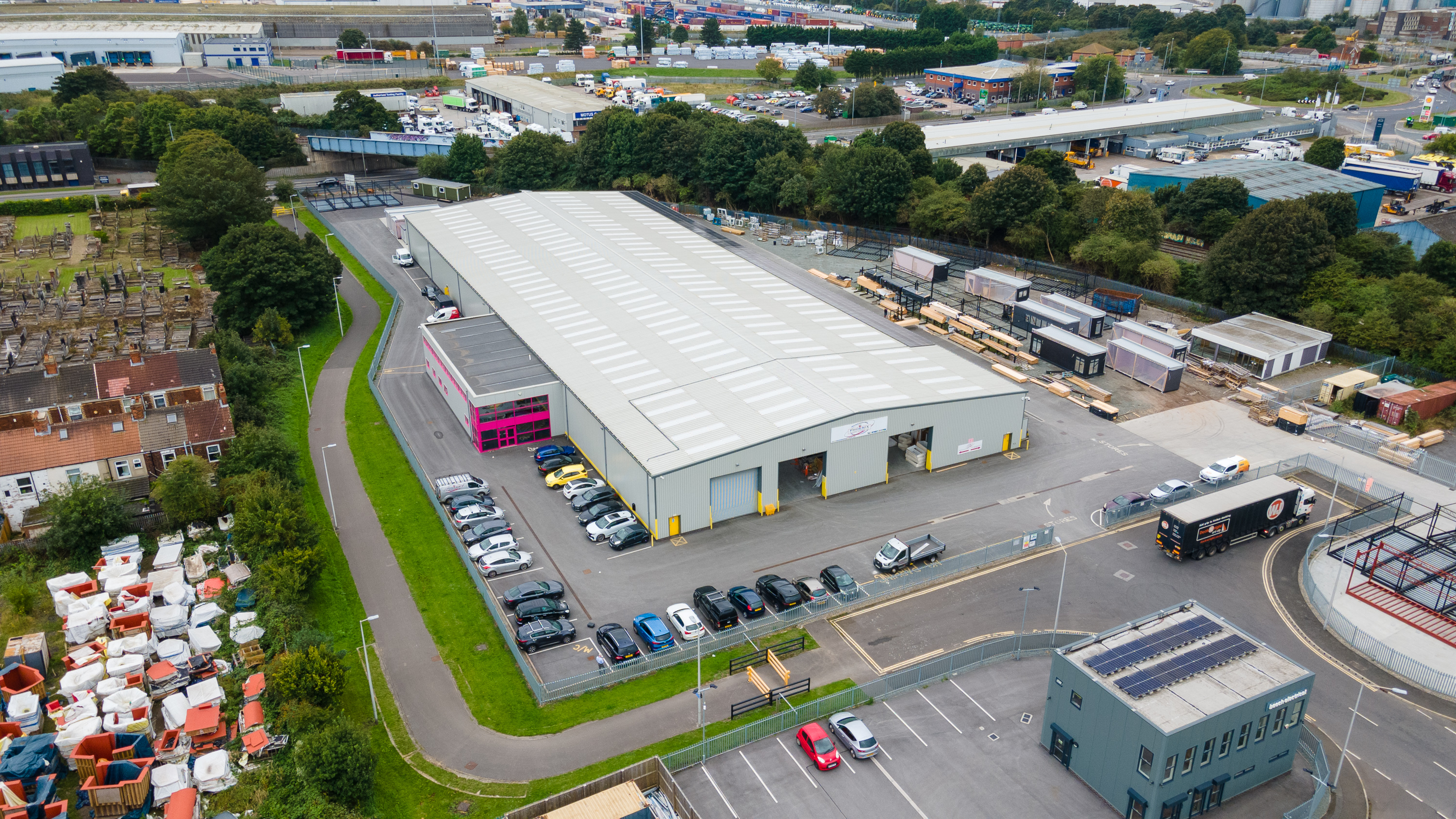 A large warehouse with parked cars and trucks outside, surrounded by trees, residential houses, and industrial areas.