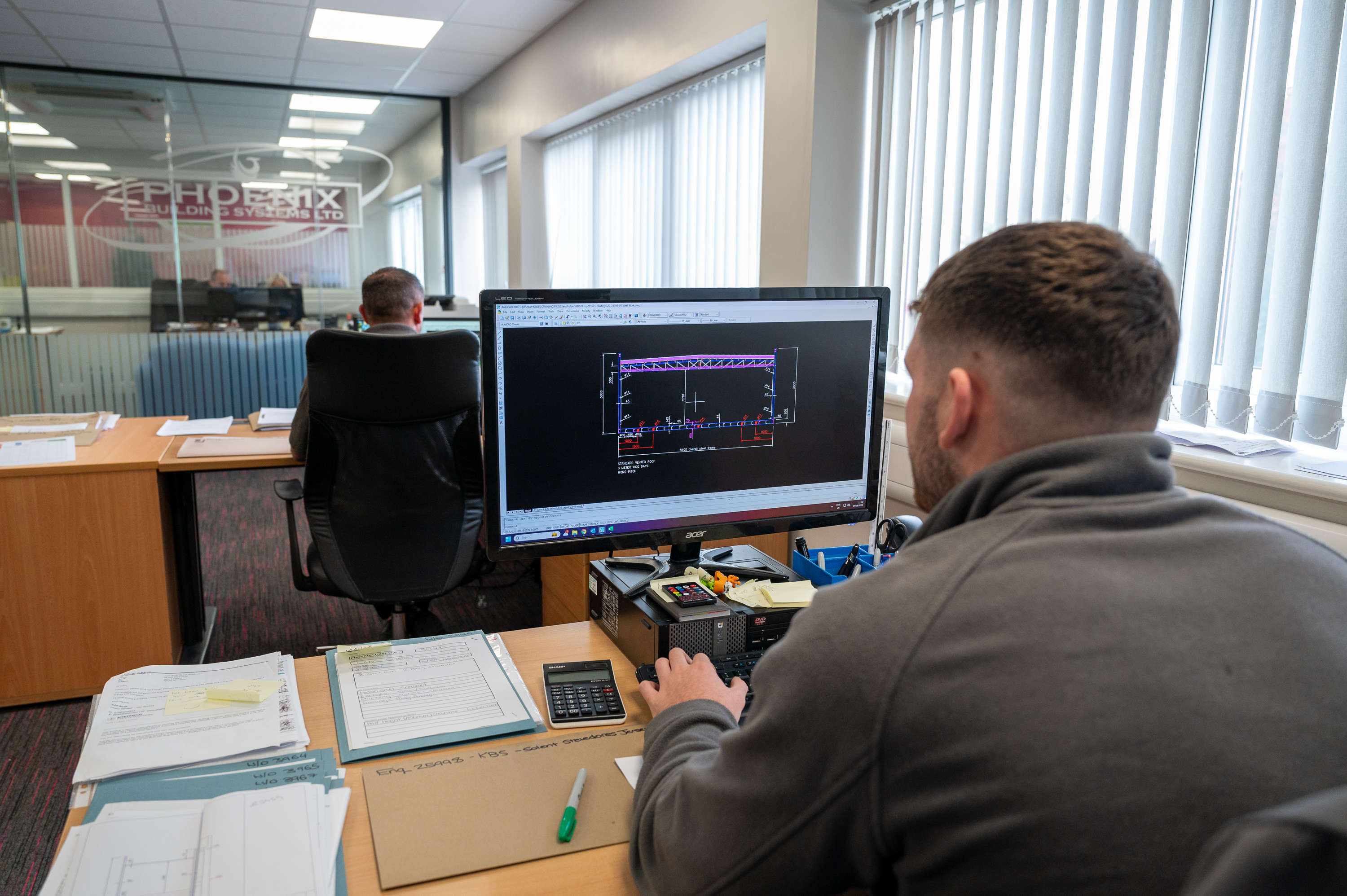 A man with tattoos is sitting at an office desk, working on a computer while holding papers. He wears a grey polo with a "PHOENIX TECHNOLOGY GROUP" logo. The office has a modern design with large windows and blinds.